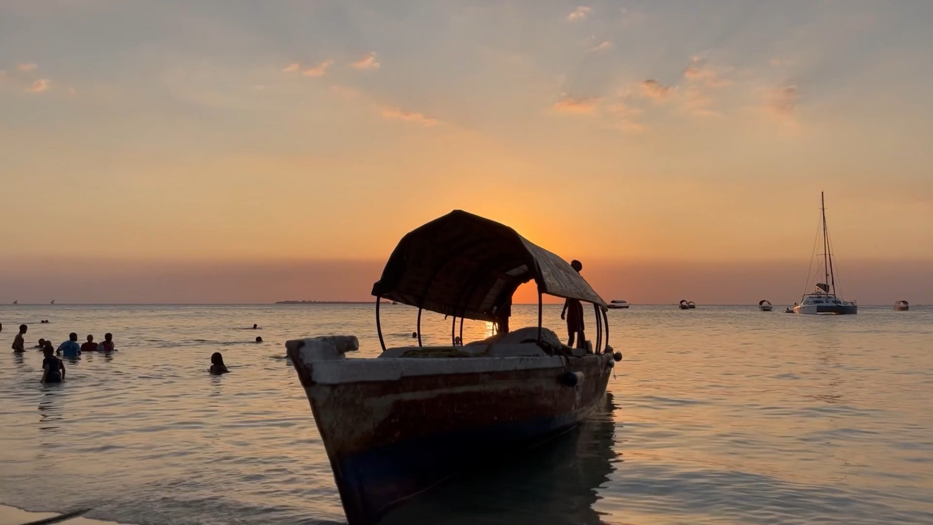 A traditional wooden dhow resting in calm water at sunset while people swim nearby